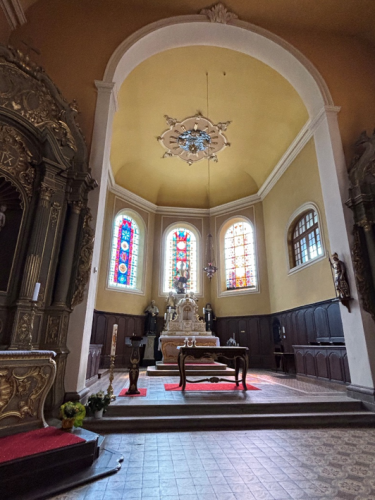 Foto eines Kircheninnenraums mit Altar, bunten Glasfenstern und kunstvoll verzierten Wänden. Helle Decke mit dekorativem Kronleuchter kontrastiert zu dunklem Holz und Mosaikboden.