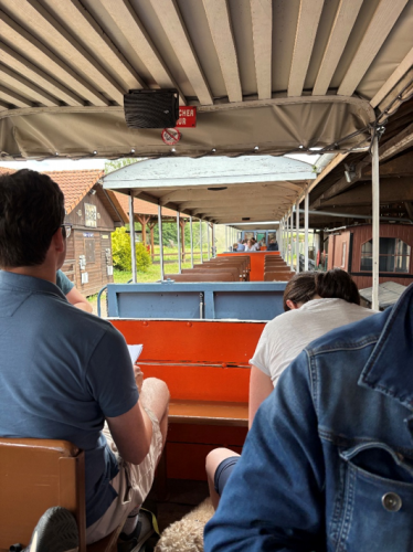 Fotografie zeigt Innenansicht eines offenen Zugabteils mit mehreren sitzenden Fahrgästen, die auf die vorbeiziehende Landschaft blicken. Orangefarbene Sitze, ein beiges Dach und grüne Bäume sowie Gebäude außerhalb des Zuges.