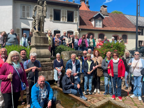 Gruppenfoto von etwa 30 Personen vor einem historischen Brunnen mit einer Statue in einem Dorf mit Fachwerkhäusern und blauem Himmel.